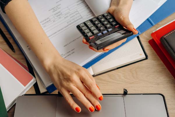 Woman making calculations by hand on documentation inside a blue folder while she moves the mouse on her laptop.