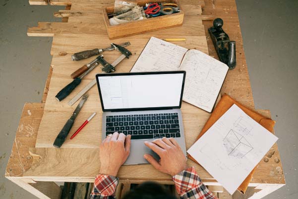 Overhead shot of a man in plaid working at a laptop surrounded by blueprints and carpentry tools.