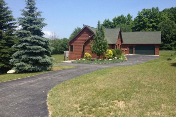 Paved driveway with tall blue spruce pines on the left side leading to a log cabin-style house with a two-car garage and decorative bushes.