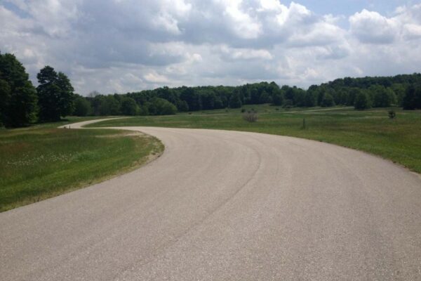 Paved road winding through a wide grass pasture with thick hardwood forests on the horizon.