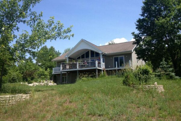 View of the back of a beige house with a covered back patio surrounded by low bushes and hardwood trees.