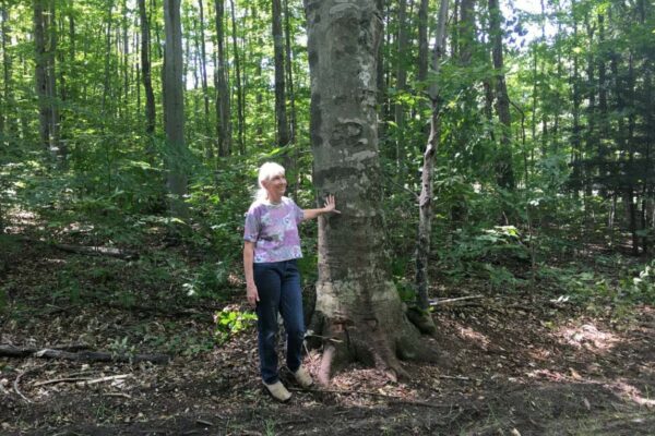 Older woman leaning against a large hardwood tree in the woods next to a dirt road.
