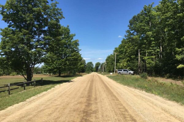 Straight dirt road with a thick forest on the right side and a wooden fence with a few large hardwood trees on the left side.