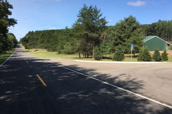 Paved two-lane road running past the entrance to the Big Sky property development with a glimpse of a green polebuilding on the right.