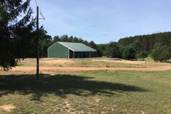 Large green polebuilding with a covered porch set back on a lot with pine trees behind it.