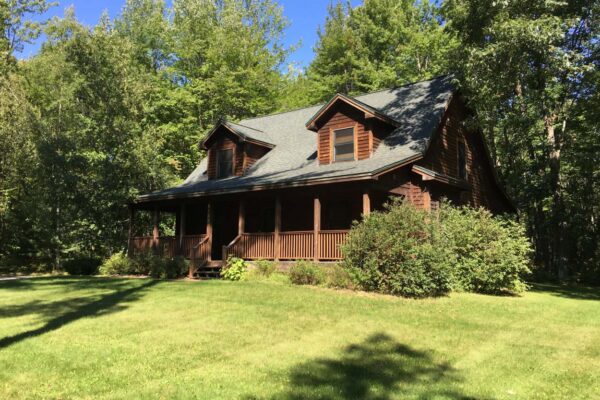 Quaint Cape Cod-style log home with covered porch surrounded by tall hardwood shade trees and a large manicured lawn.