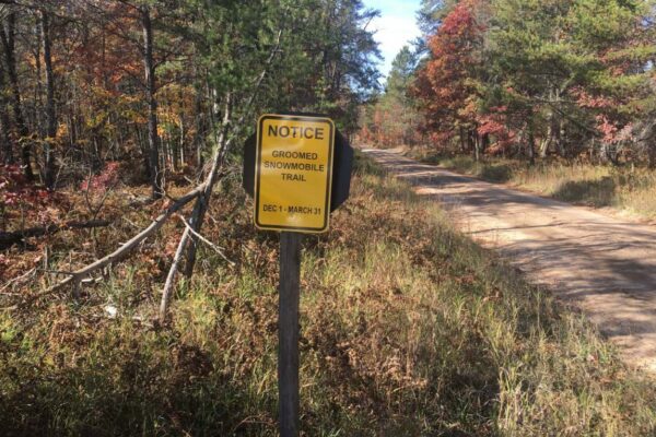 Dirt road lined with trees with a yellow sign marking the road as a groomed snowmobile trail.