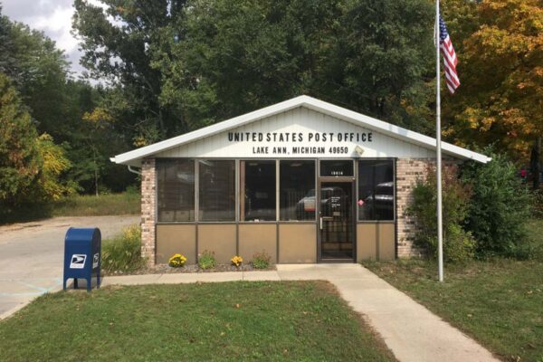 The Lake Ann post office building with an American flag on the right and a blue post office box on the left.