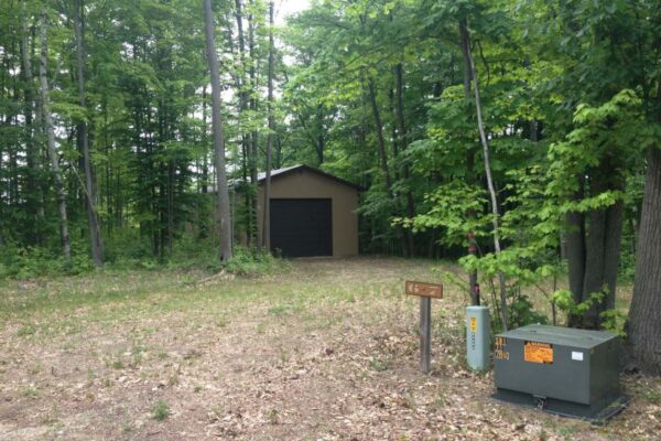 Clearing with an electrical box on the right and a beige polebuilding nestled in the trees on the left.