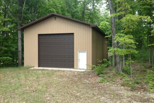 Large beige polebuilding with a dark brown door surrounded by tall hardwood trees.