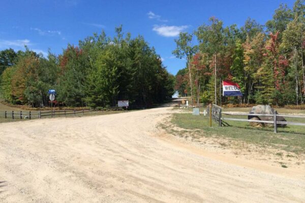 Dirt road leading into the Crofton Ridge residential property development with wooden fences on either side and a large Welcome flag on the right fence posts.