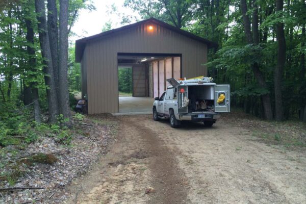 White company work truck parked at the door of a beige polebuilding in the woods.