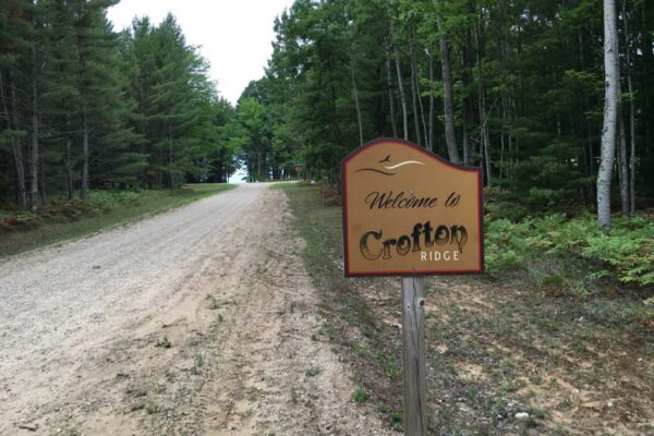 Dirt road with a Welcome to Crofton Ridge sign on the right and tall hardwood trees lining both sides.